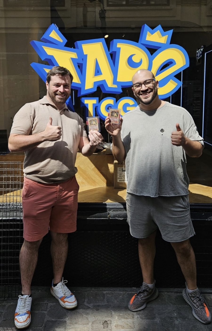 Deux hommes souriants devant la vitrine de la boutique Jade TCG à Bruxelles, pouce levé, tenant chacun une carte Dracaufeu sous pochette.